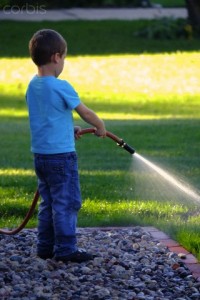 A four year old boy squirts water from a garden hose in the backyard of a home.