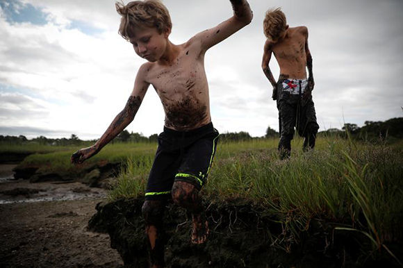 A boy leaps from the edge of a marsh while they enjoy playing in a mud bog at low tide in Barnstable Harbor on Cape Cod, Massachusetts, August 11, 2017. Photo by Mike Segar